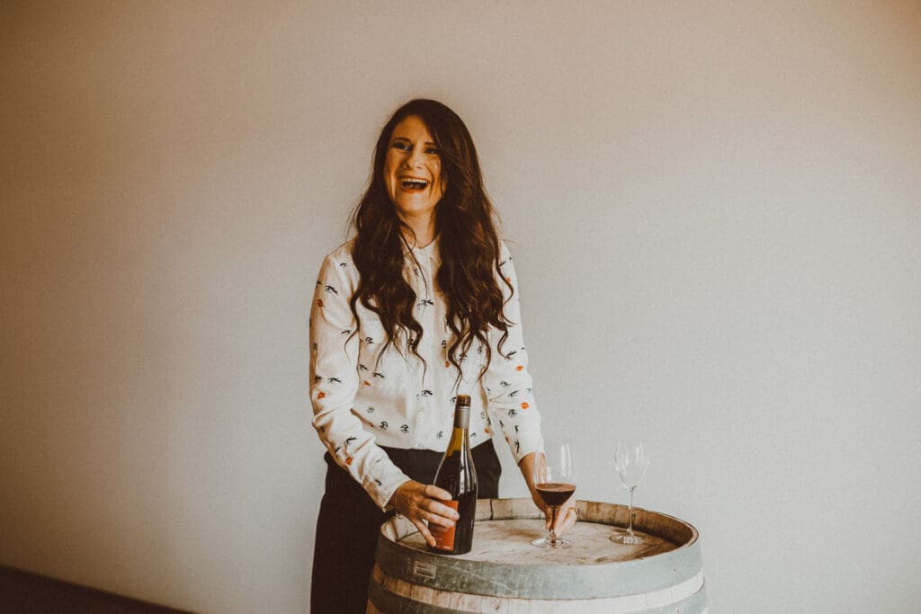 A woman with long brown hair smiles and laughs as she pours a glass of red wine, resting it on top of an old barrel.